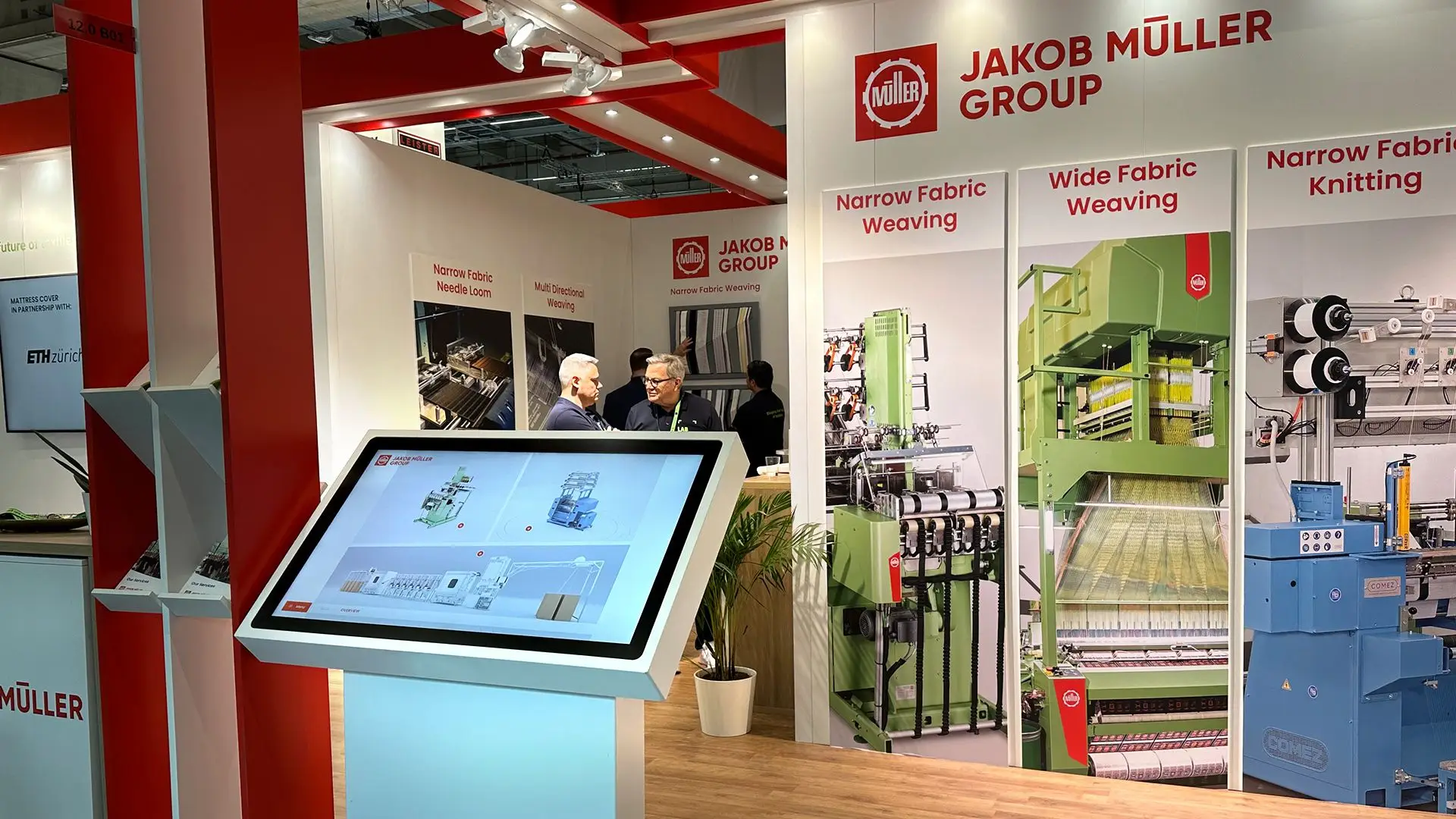 Jakob-Müller-Messestand auf der Techtextil 2026 mit Touchscreen-Terminal, Maschinenwänden und Besuchern im Gespräch